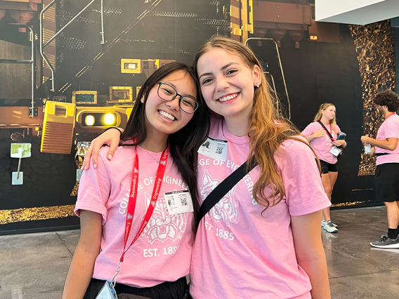 Two women in pink shirts stand in front of a mural picturing a caterpillar truck.