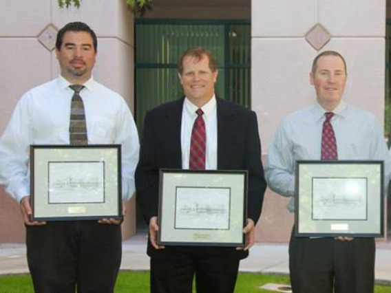 Four men in dress shirts and ties stand in a line, holding certificates in frames