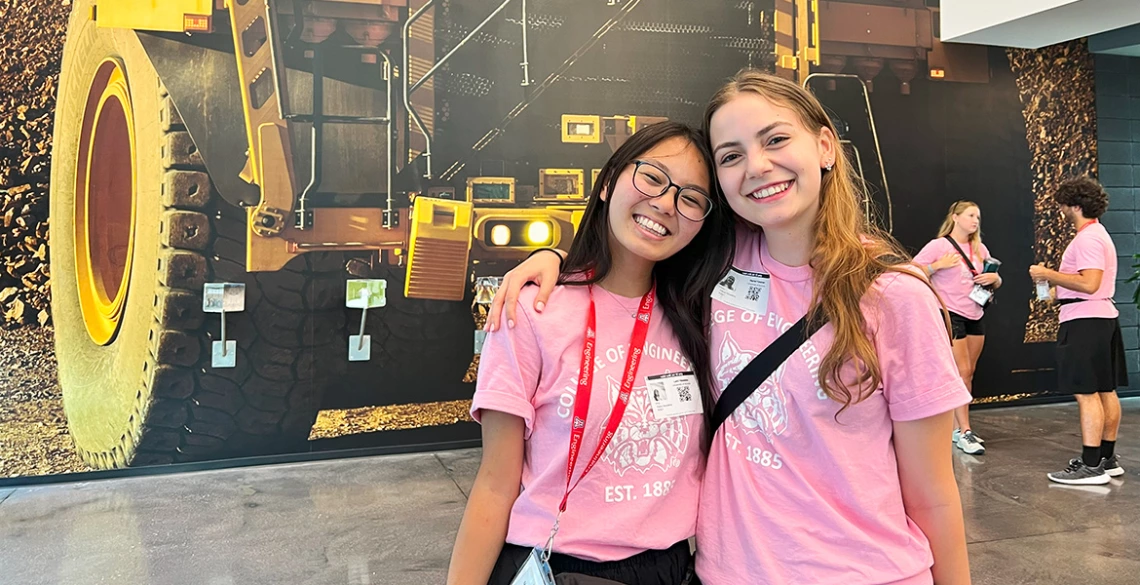 Two women in pink shirts stand in front of a mural picturing a caterpillar truck.