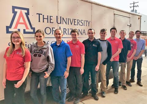 A male professor stands in line behind two female students and in front of seven male students before a white trailer bearing the University of Arizona Logo