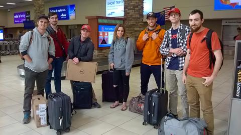 A group of students pose with their luggage in an airport with information screens in the background