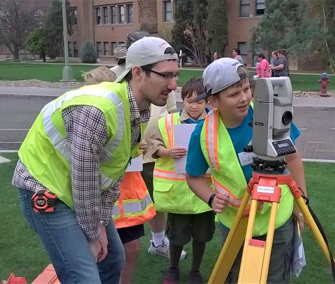 A man in a baseball cap and a yellow safety vest stands to the side of a boy, also wearing a yellow safety vest, as he peers through a theodolite