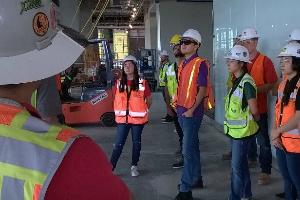 A man wearing a white hardhat and a yellow safety vest stands in the foreground and addresses a group of students who are also wearing safety gear in a partially constructed building. 