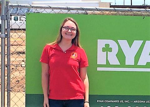 A woman wearing a red polo shirt stands before a fence and a green sign that bears a logo for Ryan Companies