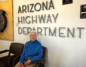 An older woman with white hair and a blue sweater sits in a chair in front of a sign that reads Arizona Highway Department.