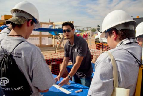 A student in a blue shirt and sunglasses stands at a table, explaining instructions to three younger students wearing hardhats and gray shirts.