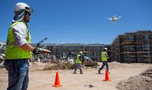 A man in a hard hat and reflective vest holds a control panel while a drone flies in front of him at a constructino site