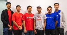 Six Cambodian students wearing UA red and blue shirts stand in front of a white wall