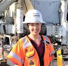 A woman in a white hardhat and an orange safety vest stands in front of construction equipment