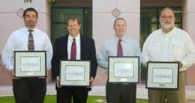 Four men in dress shirts and ties stand in a line, holding certificates in frames