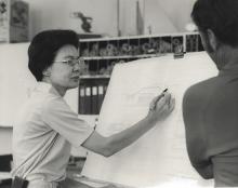 Black and white photo of a woman sketching an architectural drawing while someone else looks over her shoulder