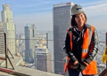 Woman standing in hardhat and orange safety vest on the balcony of a tall building, with skyscrapers behind her