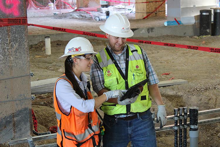 A female and male engineering student, both wearing safety gear, consult a clipboard while standing in a construction area