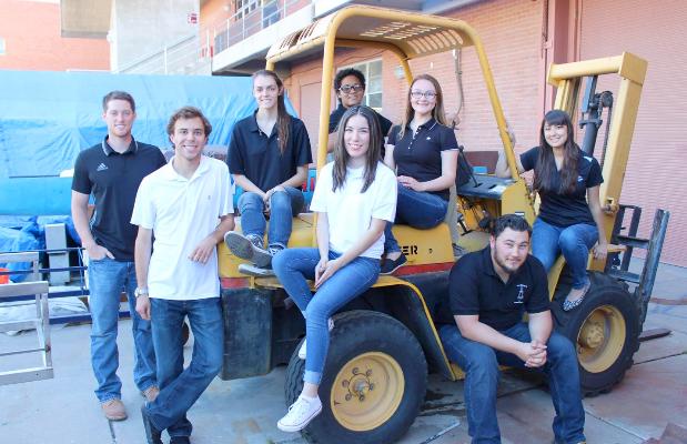 Four female and five male students sit on and lean against a yellow construction forklift
