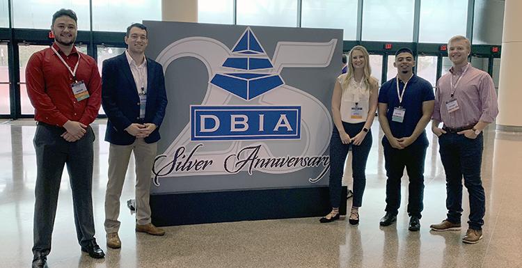 Four male students and one female student standing either side of a large poster that reads "DBIA Silver Anniversary"