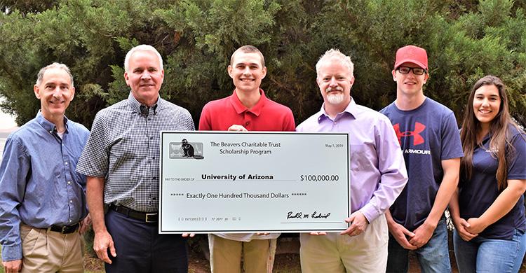 A group of six people stand in front of a tree holding an oversized check that reads "The Beavers Charitable Trust Scholarship Program." The check is made out to the University of Arizona in the amount of $100,000.