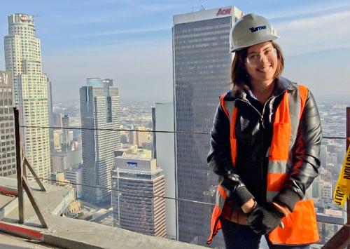 Woman standing in hardhat and orange safety vest on the balcony of a tall building, with skyscrapers behind her
