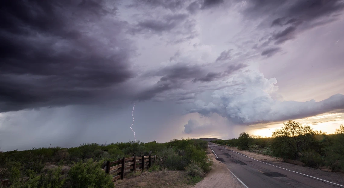 A two-lane road leads toward the horizon through a field of lush Southwestern plant life as storm clouds gather overhead. In the distance, rain and lightning can be seen.