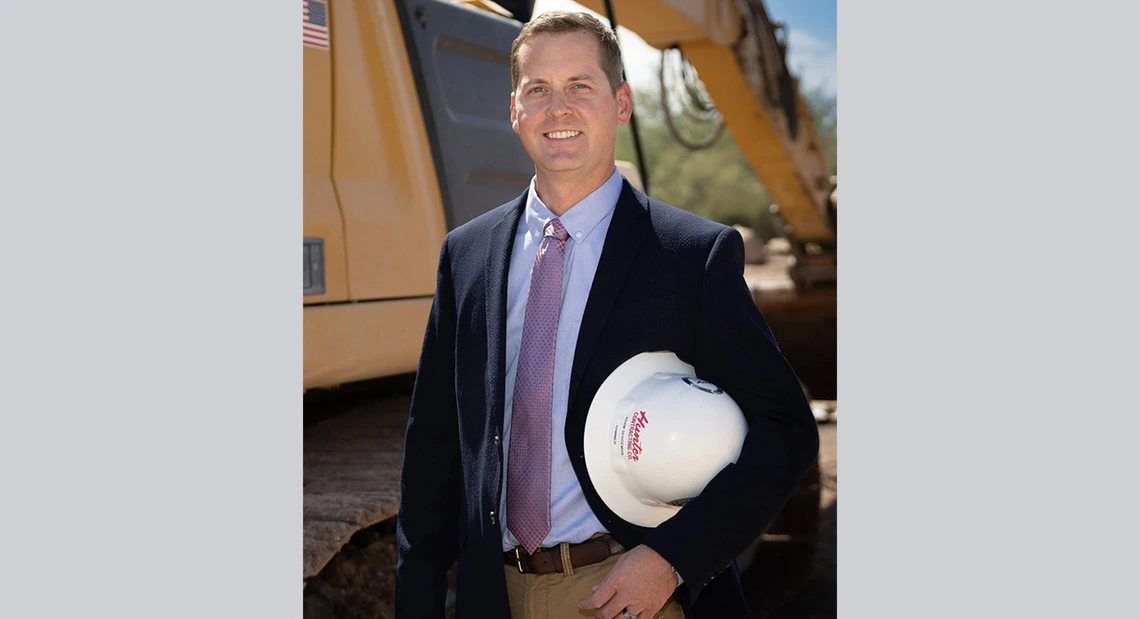 Man in coat in tie stands holding a hard hat in front of construction equipment.