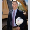 Man in coat in tie stands holding a hard hat in front of construction equipment.